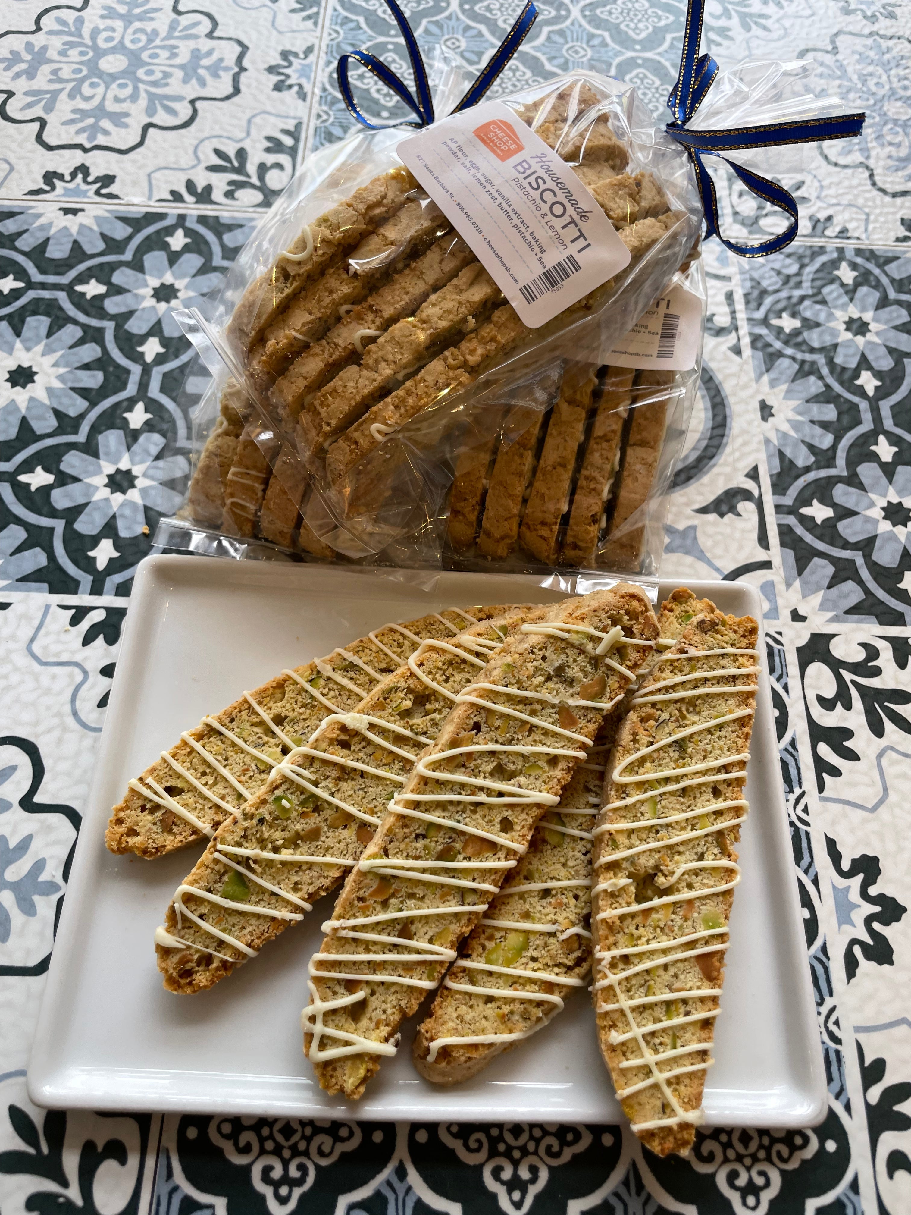 3 bags of 5 slices of pistachio biscotti, with 5 pieces laid out on a plate with slight white chocolate drizzle