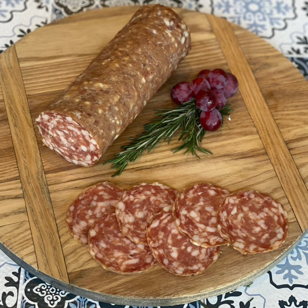 6 slices of calabrese, on a wooden circle cutting board, next to half a log of calabrese, with bunch of grapes and rosemary twig