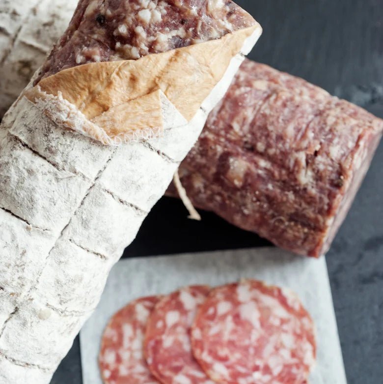 Two whole pieces of salami with skin partially peeled and some sliced Finocchiona in background