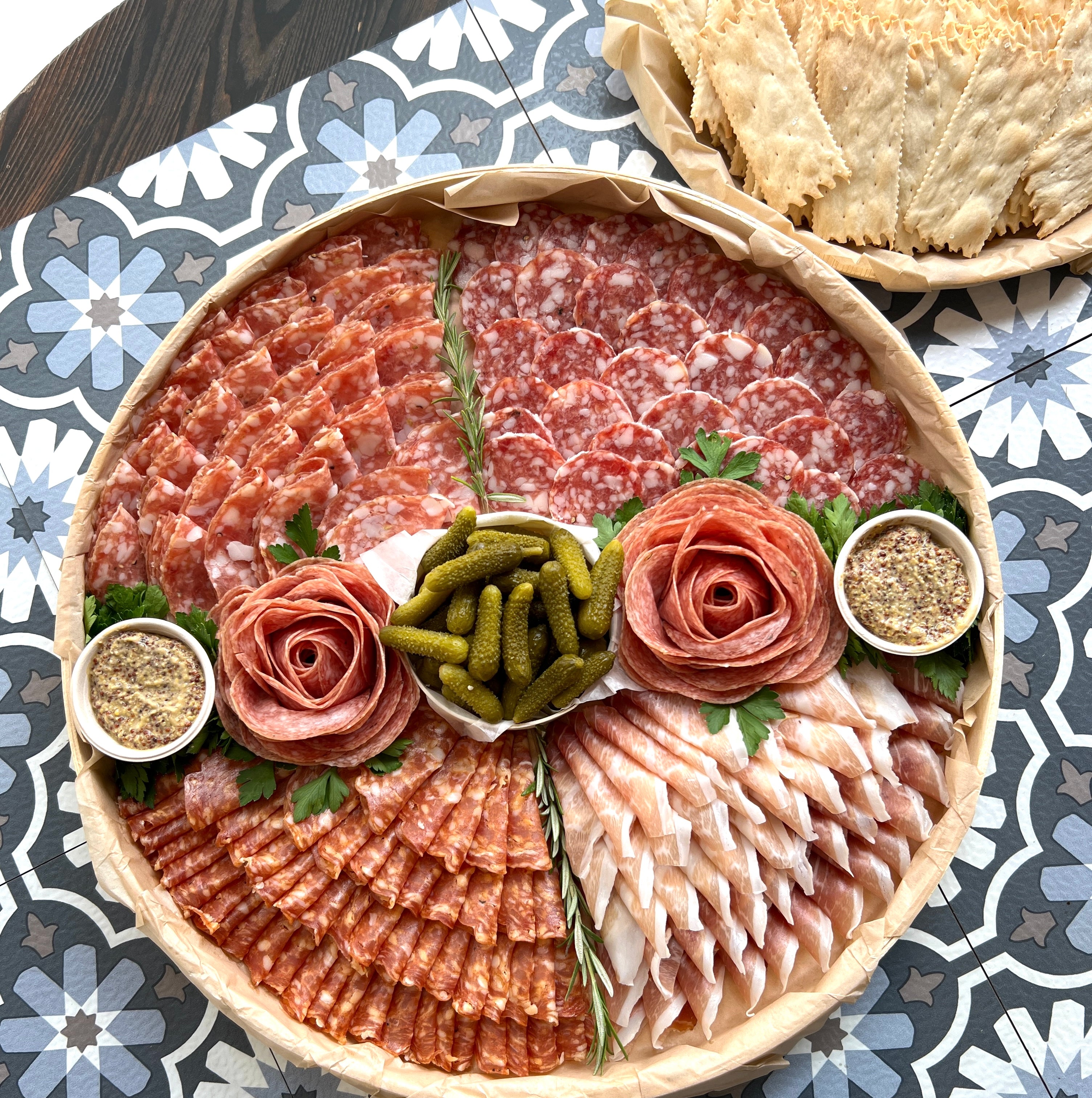 Our large Salumi platter featuring two salami roses surrounded by four other types of folded cured meat and cornichons in the center.  Small mustards are on also on the platter and a side plate of panzanella crackers in the background.