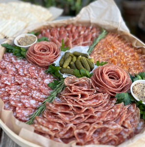 Our large Salumi platter featuring two salami roses surrounded by four other types of folded cured meat and cornichons in the center.  Small mustards are on also on the platter and a side plate of panzanella crackers in the background.