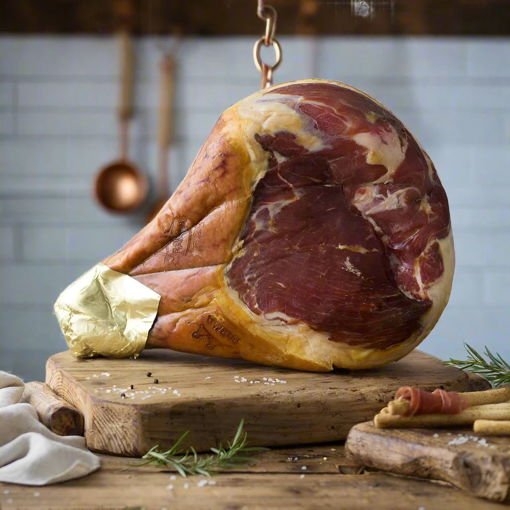 Whole leg of prosciutto on its side sitting on a thick board with some rosemary in a kitchen setting