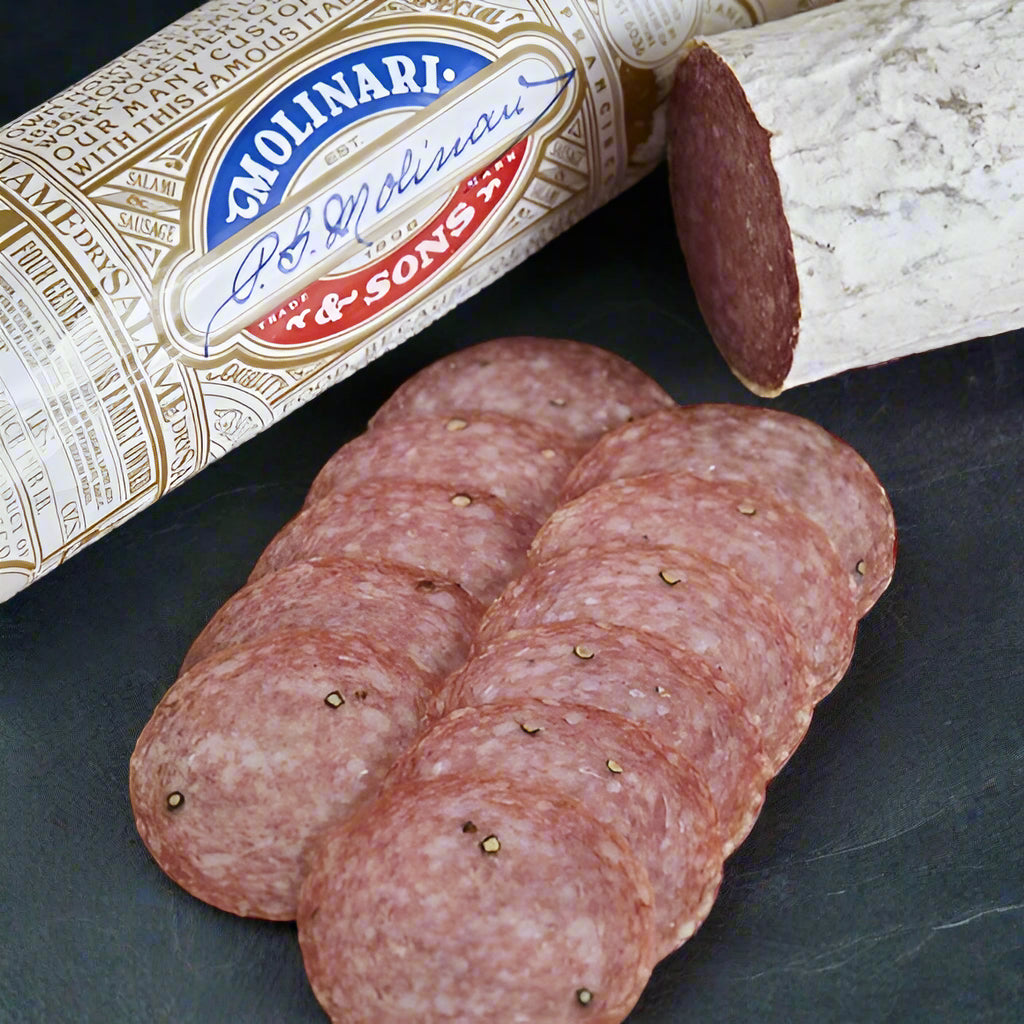 2 sticks of Italian Dry salami, one fully wrapped with the Molinari logo in focus.  Sliced pieces of salami in the foreground.